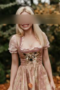 SP Escort Agency Munich, Carolin in a rose Dirndl. Her blond hair is falling over her shoulder while she stands in a garden.