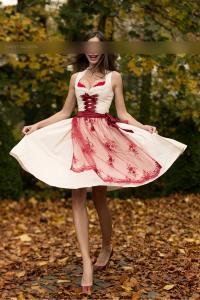 SP Escort Agency Munich, Jessica in a beige-red Dirndl with highheels. Shes swinging her skirt whilst smiling, in a garden.