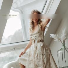 SP Escort Agency Munich, Laura in a golden Dirndl, standing with one leg on a couch. She's smiling and has a hand in her hair.